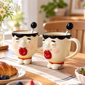 Puede incluir: Dos tazas de cerámica color crema con forma de rostros humanos, cabello negro, narices prominentes y labios rojos fruncidos. Cada taza tiene un asa lateral y una cuchara pequeña con punta negra. La base de cada taza se asemeja a unos pies.