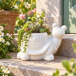 May include: A white ceramic turtle-shaped planter featuring a glossy finish and a shell designed to hold plants. The planter is filled with a variety of green and pink succulents, including a trailing string of pearls plant cascading over the side.