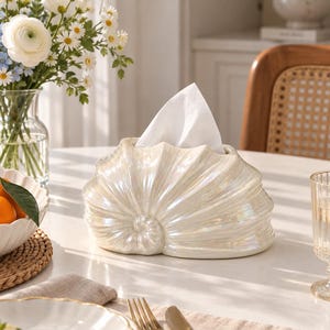 Puede incluir: Una funda para caja de pañuelos de color blanco perla con forma de concha marina grande, con un centro en espiral y textura acanalada. El soporte decorativo presenta una amplia abertura en la parte superior, de la que sobresale un pañuelo blanco.