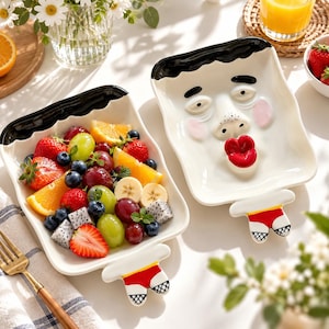 Puede incluir: Dos platos rectangulares de cerámica con un rostro humano esculpido, cabello negro, mejillas rosadas y labios rojos prominentes. Un plato está lleno de fruta fresca, mientras que el otro plato está vacío.