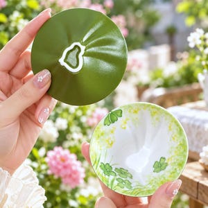 Könnte beinhalten: Eine Keramik-Teetasse mit passendem Deckel, die ein grünes Äußeres mit einem weißen Mittelakzent auf dem Deckel aufweist. Das Innere der Tasse zeigt eine weiße Basis mit einem zarten grünen Kleemuster und sanften gelben Blumenakzenten am Rand.