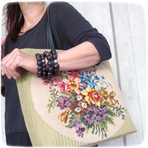 Peut inclure: Sac fourre-tout en velours côtelé beige avec un grand motif floral brodé multicolore. Le sac a une bandoulière en cuir noir et est porté par une personne vêtue d'un haut noir et de bracelets de perles en bois foncé.