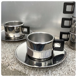 May include: Two stainless steel coffee cups with black handles and matching saucers. The cups are reflective and have a modern design.