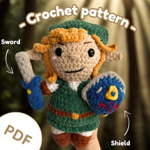 Peut inclure: Poupée crochetée d'un personnage de jeu vidéo, tenant une épée et un bouclier. La poupée porte des vêtements verts, des cheveux blonds et un chapeau vert. Le bouclier est bleu avec un emblème jaune. Le texte "Crochet pattern" et "PDF" est inclus.