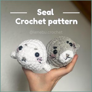 Könnte beinhalten: Zwei gehäkelte Seehund-Plüschtiere, eines weiß und eines grau, werden in einer Hand gehalten. Die Seehunde haben schwarze Augen, rosa Wangen und gestickte Details. Der Text auf dem Bild lautet "Seal Crochet pattern" und der Instagram-Handle "@lenebu.crochet."