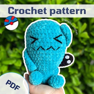 Op de afbeelding: Een turquoise gehaakt pluche speelgoed met zwarte gezichtskenmerken en een klein zwart aanhangsel. De afbeelding bevat de tekst "Crochet pattern" en "PDF". De achtergrond is een vage groene blad.