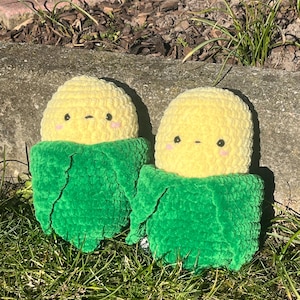 Könnte beinhalten: Zwei gehäkelte Mais-Plüschtiere mit grünen Hüllen und gelben Körnern. Die Plüschtiere haben gestickte Augen und einen kleinen Mund.