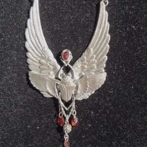 Op de afbeelding: Een zilveren ketting met een gevleugelde scarabee-hanger en rode edelsteenaccenten. De hanger is gedetailleerd en hangt aan een ketting met rode kralen en bruine stenen kralen.