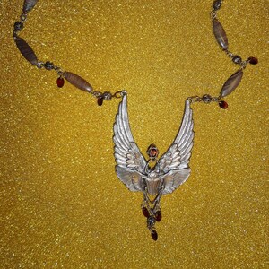 Op de afbeelding: Een zilveren ketting met een gevleugelde scarabee-hanger en rode edelsteenaccenten. De ketting is gemaakt van bruine en zilveren kralen.