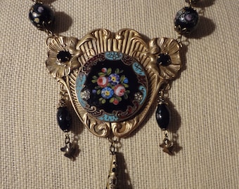 May include: A gold-tone necklace with a black enamel pendant featuring a floral design. The pendant is surrounded by a decorative frame and hangs from a chain with black beads.