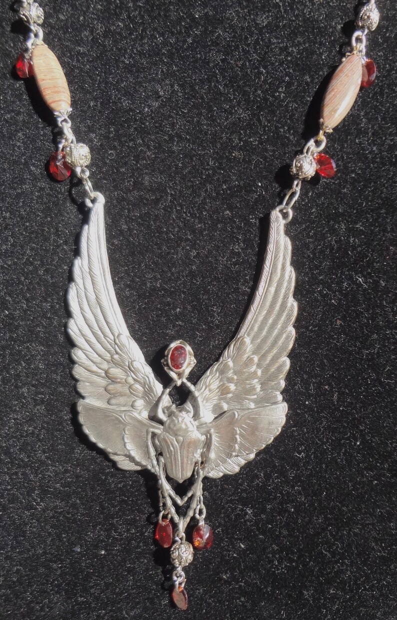 Op de afbeelding: Een zilveren ketting met een gevleugelde scarabee-hanger en rode edelsteenaccenten. De hanger is gedetailleerd en hangt aan een ketting met rode kralen en bruine stenen kralen.