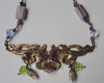 May include: A vintage necklace with a bronze-coloured filigree design. The necklace features lavender-coloured rectangular beads, purple beads, and clear iridescent beads. It is adorned with translucent purple flowers, green leaves, and pink and green buds.