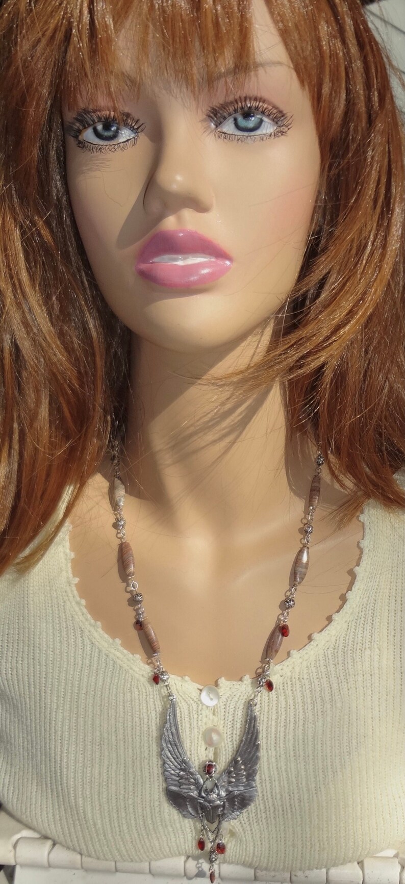 Op de afbeelding: Een close-up van een vrouw die een witte trui draagt met een zilveren gevederde hangerketting met rode kralen. De ketting heeft een gedetailleerd gevederd ontwerp met een hart in het midden.