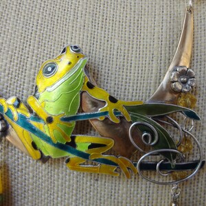 Könnte beinhalten: Handgefertigte Halskette mit einem bunten Frosch-Anhänger. Der Frosch ist gelb und grün mit schwarzen Akzenten, platziert auf einem gebogenen Metallrahmen mit floralen und Blattdetails. Einzigartiges, künstlerisches Design.