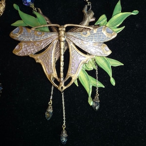 May include: A detailed dragonfly pendant necklace with a bronze-colored body and lavender wings, accented with green leaves and dangling blue crystal beads. The necklace is set against a black background.
