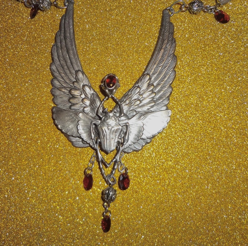 Op de afbeelding: Zilveren ketting met een keverontwerp en rode edelsteenaccenten. De kever heeft vleugels en is omgeven door een ketting met rode edelstenen.