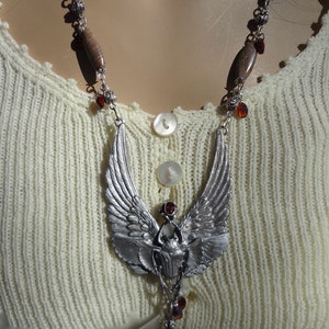 Op de afbeelding: Een zilveren ketting met een gevleugelde scarabee-hanger en bruine houten kralen. De ketting ligt op een witte gebreide trui.