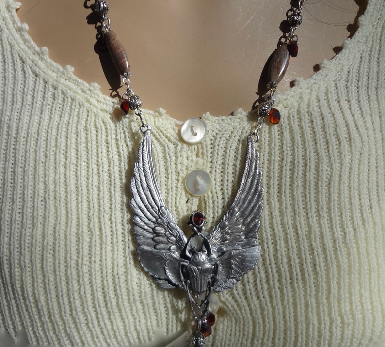 Op de afbeelding: Een zilveren ketting met een gevleugelde scarabee-hanger en bruine houten kralen. De ketting ligt op een witte gebreide trui.