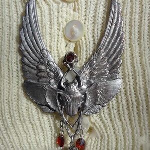 Op de afbeelding: Een zilveren ketting met een gevleugelde scarabee-hanger en rode edelsteenaccenten. De hanger hangt aan een ketting met meerdere rode edelsteendruppels.