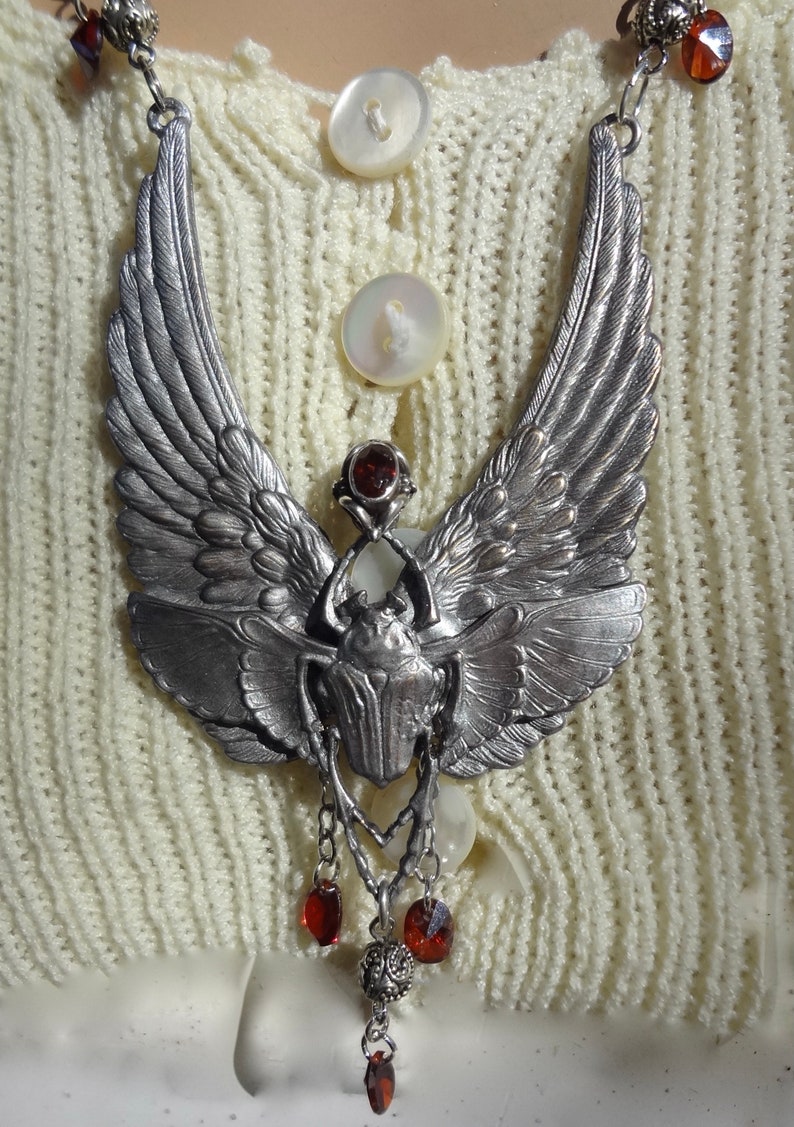 Op de afbeelding: Een zilveren ketting met een gevleugelde scarabee-hanger en rode edelsteenaccenten. De hanger hangt aan een ketting met meerdere rode edelsteendruppels.