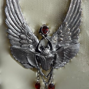 Op de afbeelding: Een zilveren ketting met een hanger in de vorm van een gevleugelde scarabee met een rode edelsteen. De hanger hangt aan een ketting met drie rode edelsteen accenten.