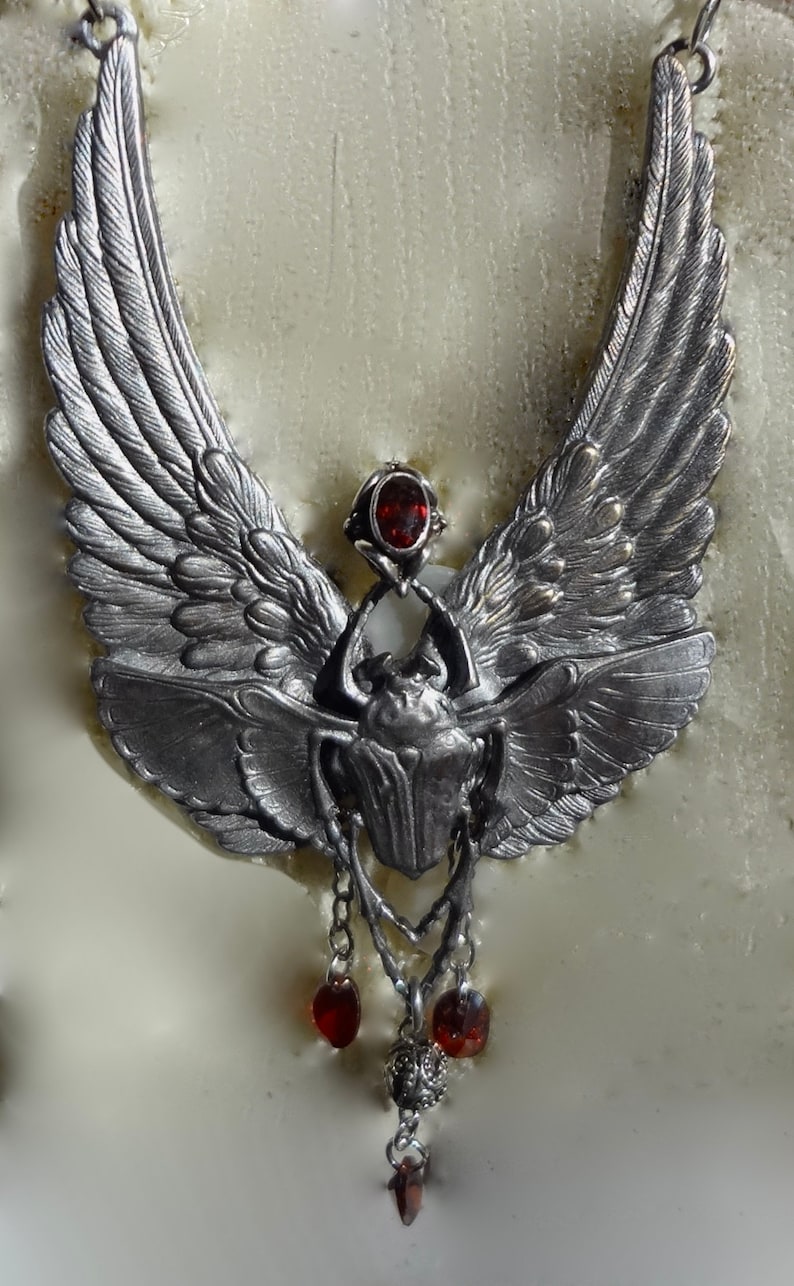 Op de afbeelding: Een zilveren ketting met een hanger in de vorm van een gevleugelde scarabee met een rode edelsteen. De hanger hangt aan een ketting met drie rode edelsteen accenten.