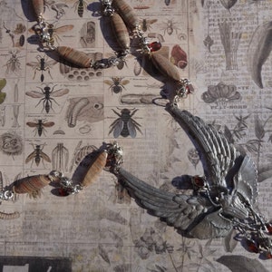 Op de afbeelding: Een zilveren ketting met een gevleugelde hanger en rode edelsteenaccenten. De ketting is gemaakt met bruine en witte gepatenteerde kralen en zilveren kettingschakels. De hanger heeft een gedetailleerd vleugelontwerp en een kleine, ronde zilveren hanger.
