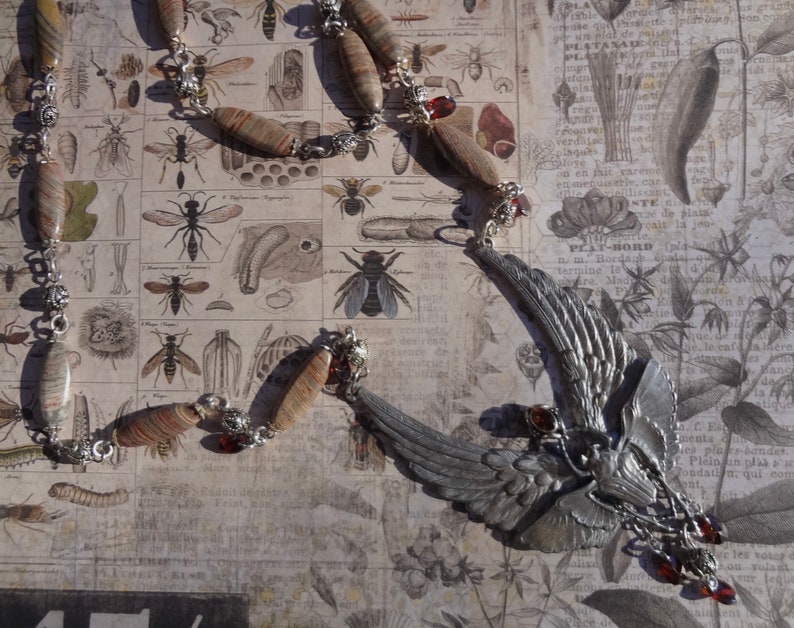 Op de afbeelding: Een zilveren ketting met een gevleugelde hanger en rode edelsteenaccenten. De ketting is gemaakt met bruine en witte gepatenteerde kralen en zilveren kettingschakels. De hanger heeft een gedetailleerd vleugelontwerp en een kleine, ronde zilveren hanger.