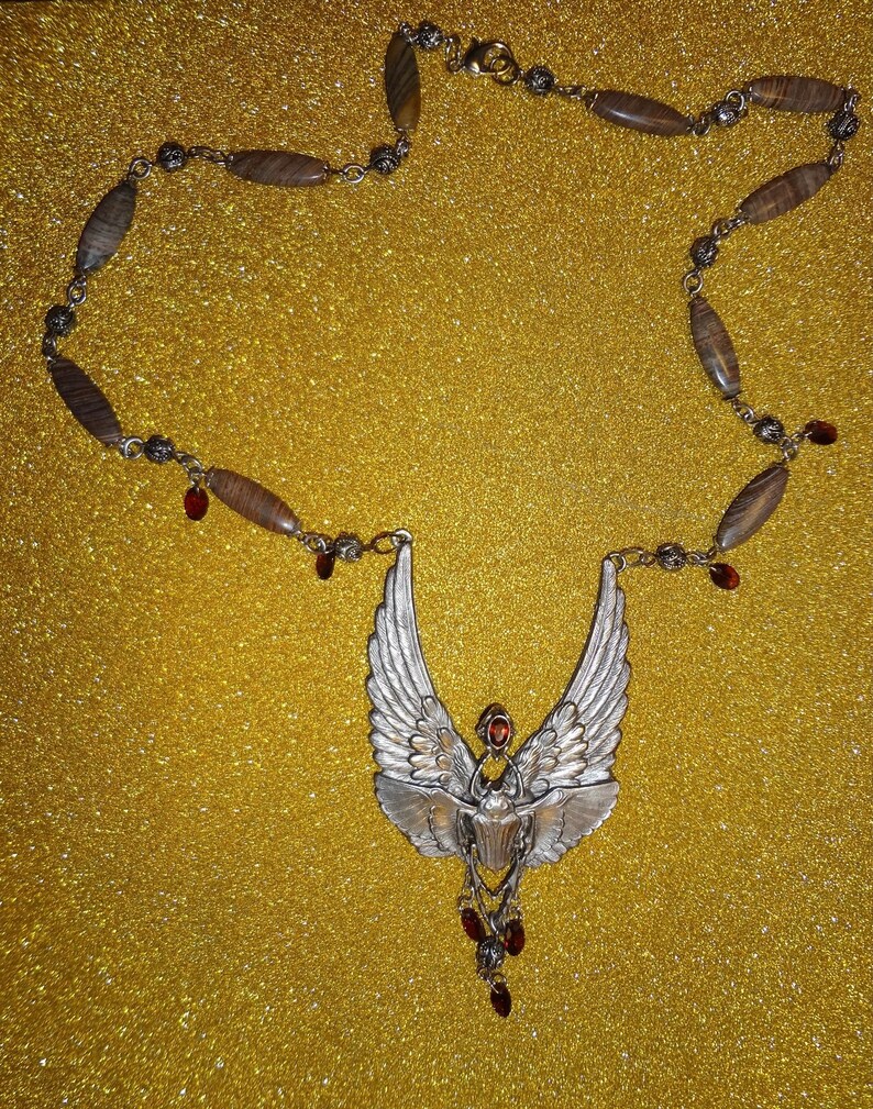 Op de afbeelding: Een zilveren ketting met een gevleugelde scarabee-hanger en rode edelsteenaccenten. De ketting is gemaakt van bruine en zilveren kralen.
