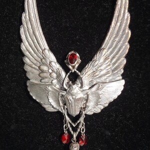 Op de afbeelding: Zilveren ketting met een gedetailleerd vleugelontwerp en een keverhanger. De hanger is versierd met een rode edelsteen en hangt aan een ketting met rode edelsteenaccenten.