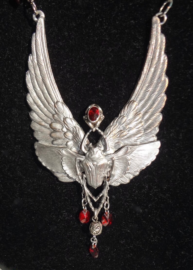 Op de afbeelding: Zilveren ketting met een gedetailleerd vleugelontwerp en een keverhanger. De hanger is versierd met een rode edelsteen en hangt aan een ketting met rode edelsteenaccenten.