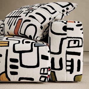 Könnte beinhalten: Nahaufnahme eines Sofa- und Kissensets mit weißem Hintergrund und einem schwarzen geometrischen Muster. Das Muster umfasst Formen und Linien mit Akzenten in Orange, Gelb, Blau und Rot. Das Sofa und das Kissen stehen auf einem hellbeigen Teppich.