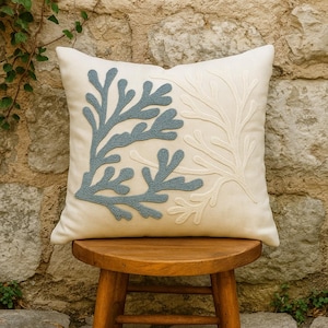 May include: Cream-coloured square cushion featuring embroidered coral reef designs in light blue and white. The cushion is placed on a small wooden stool against a stone wall, evoking a coastal theme.