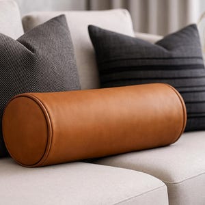 May include: A tan leather bolster cushion sits on a cream sofa, with two dark grey throw cushions either side. One cushion has a herringbone pattern, the other has horizontal stripes. The scene suggests a modern living room setting.