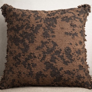 May include: Square decorative pillow with a textured, mottled design in shades of brown and dark brown. The pillow has a fringed edge, adding a rustic touch. The pillow is likely used for home decor.