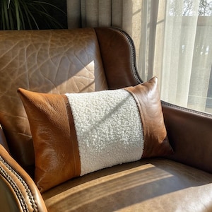 May include: A white faux fur cushion with brown leather trim sits on a brown leather armchair. The cushion is positioned in the centre of the chair, with the leather trim facing outwards.