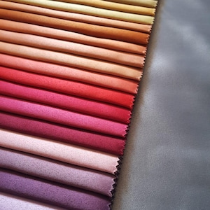 Può includere: Un campionario di tessuti con una gamma di colori, che vanno dal turchese al viola scuro. I campioni sono disposti in gradiente, mostrando una varietà di sfumature. Un tessuto grigio si trova sul lato destro dell'immagine.