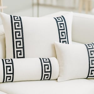 May include: Four ivory decorative pillows with a black Greek key design. The set includes two square pillows, a rectangular pillow, and a cylindrical bolster pillow, all arranged on a white sofa.