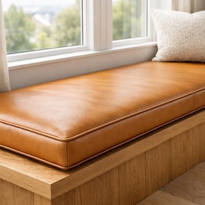 May include: A rectangular, caramel-colored leather cushion sits atop a wooden bench, with a window and curtains in the background. The cushion is approximately 60 inches long and 20 inches wide, with a stitched border. Two pillows rest on the bench.