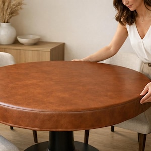 Può includere: Un tavolo rotondo in pelle marrone con base nera. Il tavolo ha un diametro di circa 122 cm. L'immagine mostra il tavolo in una stanza ben illuminata con toni neutri e una persona che lo tocca.