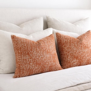 May include: Two square cushions with a rust-coloured, textured pattern rest on a white bed. The cushions are flanked by white cushions, creating a layered look. The bed is covered with white linen.