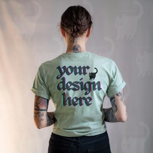 May include: A light green t-shirt with rolled-up sleeves. The back of the shirt features the text "your design here" in a stylized font with a black cat graphic. The shirt is made of a soft material.