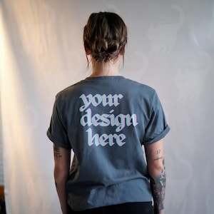 May include: A slate blue t-shirt with rolled-up sleeves. The back of the shirt features the text "your design here" in a white, 3D-style font. The person wearing the shirt has tattoos on their arm.