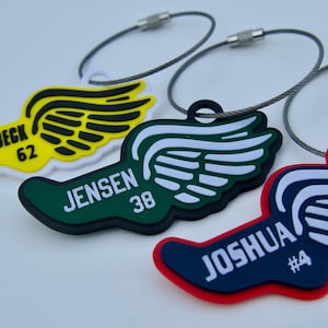 Może przedstawiać: Trzy kolorowe zawieszki do bagażu w kształcie uskrzydlonych stóp. Każda zawieszka ma inny schemat kolorów: żółty z czarnymi skrzydłami, zielony z białymi skrzydłami i granatowy z białymi skrzydłami. Każda zawieszka ma imię i numer.