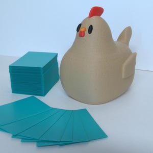 May include: A tan chicken-shaped container with a red comb, orange beak, and black eyes, positioned next to a stack of rectangular teal cards and several loose teal cards fanned out in front of the container on a flat white surface.