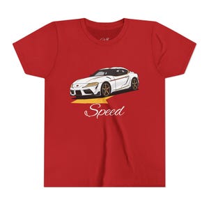 Può includere: T-shirt rossa con grafica di auto sportiva bianca, dettagli dorati e la parola "Speed" in corsivo. L'auto è su una freccia gialla, che suggerisce velocità. Un design per gli appassionati di auto.