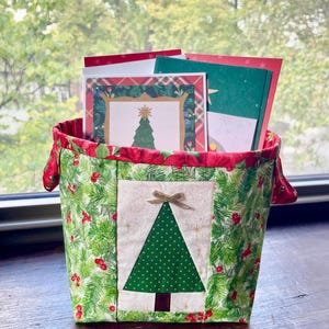 Peut inclure: Un panier en tissu rempli de cartes de Noël. Le panier est décoré d'un motif de vacances vert et rouge, avec une application de sapin de Noël sur le devant. Les cartes présentent des motifs festifs.