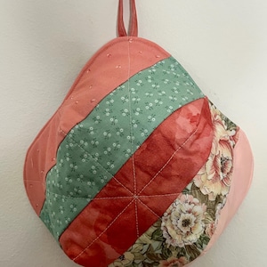 Puede incluir: Un agarrador acolchado con un lazo para colgar. El agarrador presenta un diseño de patchwork con rayas diagonales de tela coral, roja y floral. La tela floral tiene un fondo crema con flores rosas y verdes.