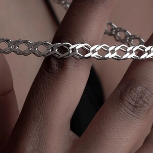 May include: A silver-toned chain necklace featuring a series of interlocking, flattened oval links. The metal surface is polished and reflective, creating a bright metallic sheen across the entire length of the chain as it curves in a loop.