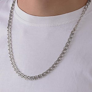 May include: A silver-toned chain necklace featuring a double-link design with a polished, reflective finish. The chain rests against a plain white fabric background, showcasing the intricate, interlocking metal links that form a continuous pattern.